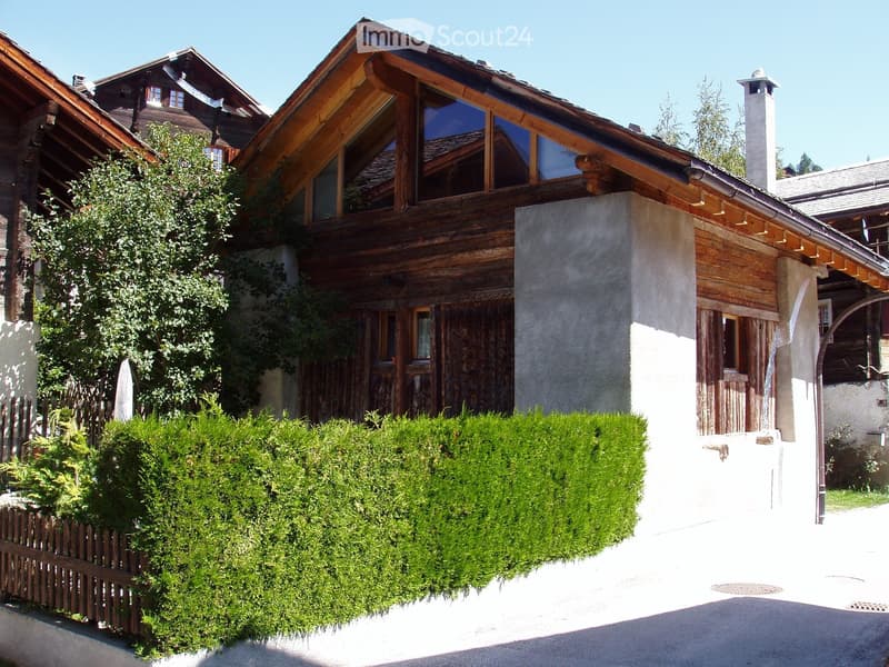 Two-story wooden house with a sloped roof, large windows, and a stone base. The house is surrounded by greenery and a wooden fence.