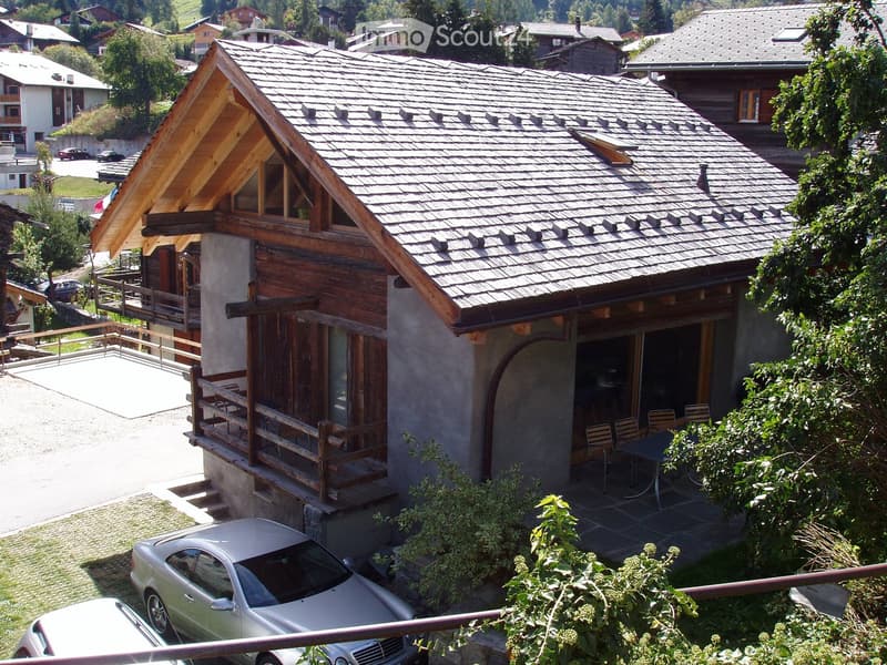 A two-story wooden house with a tiled roof, balcony, and outdoor seating area. The house is surrounded by greenery and has a parking area with a silver car.