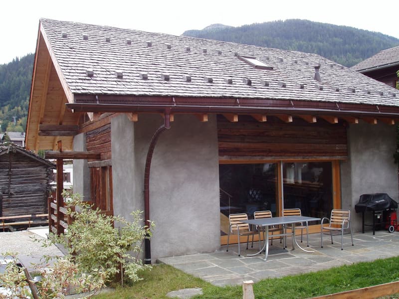 Outdoor patio area with a stone floor, a metal table and chairs, a covered entrance to the house, and a grill. The house has a wooden exterior and a tiled roof, with a mountainous backdrop.
