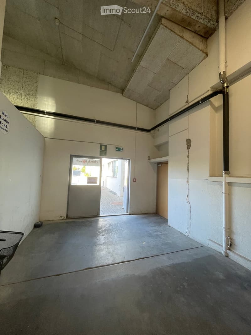 Empty hallway with concrete floor, white walls, exposed pipes, and a door leading outside.