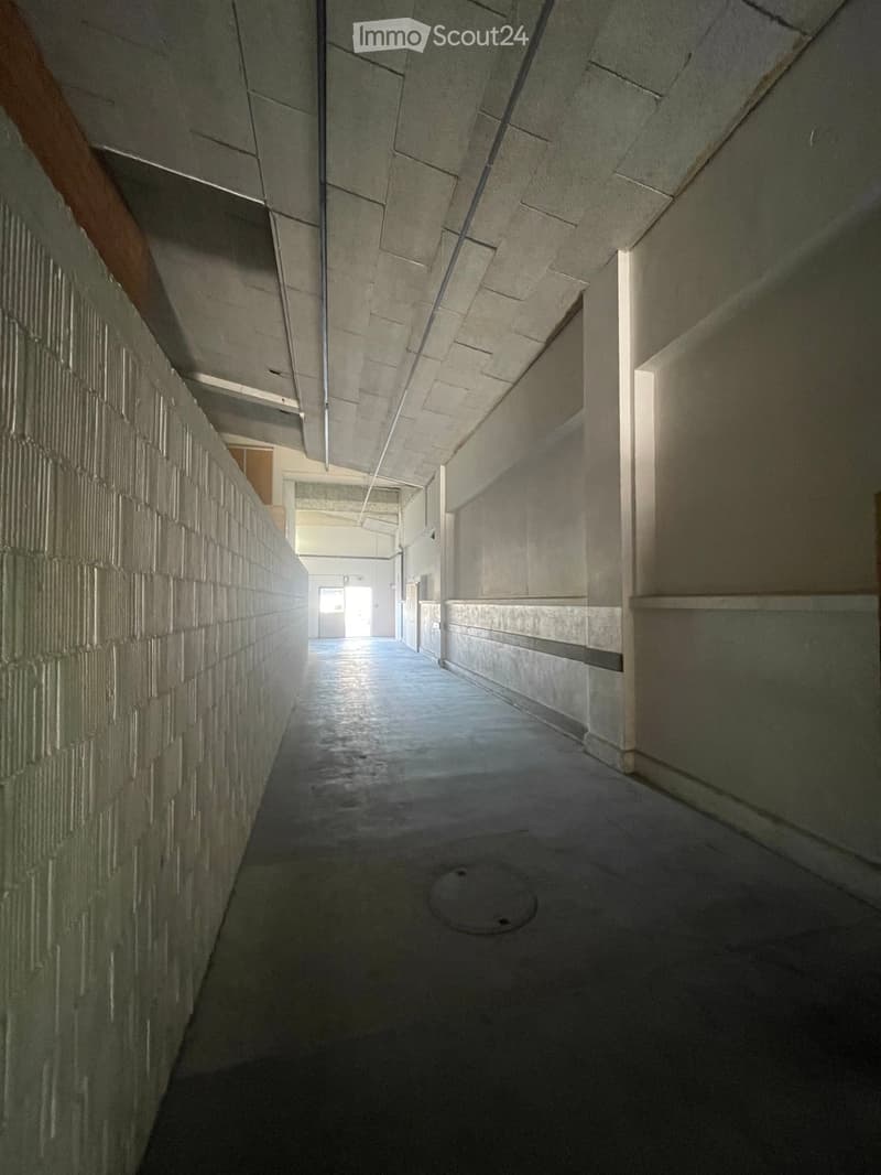 A long, narrow hallway with concrete walls and a smooth floor, featuring a metal grate in the center and a visible door at the far end.