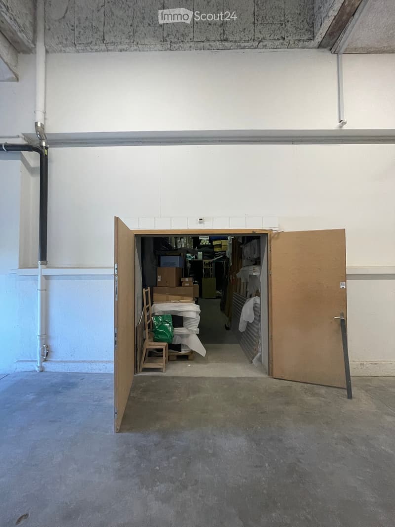 Empty room with concrete floor, white walls, and an open wooden door leading to a storage area with boxes and a chair.