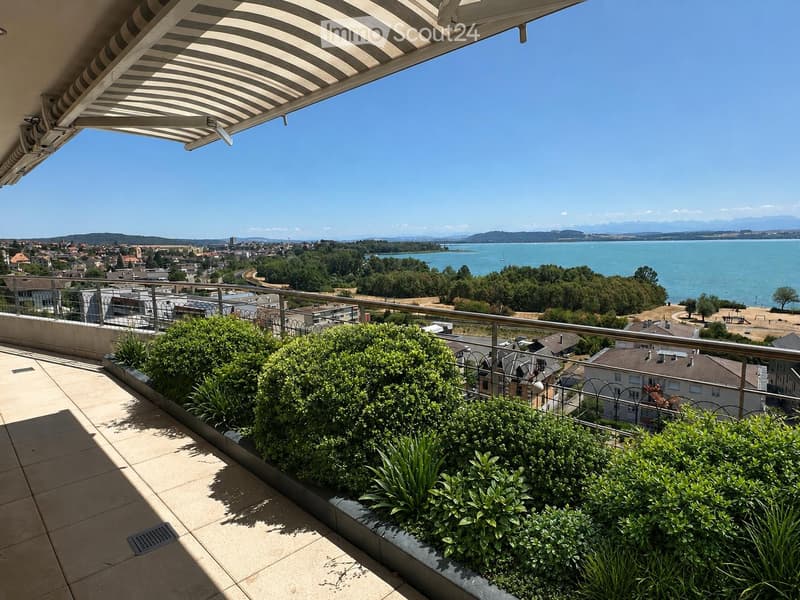 Balcony with a view of a lake and town, featuring a striped awning, metal railing, and neatly trimmed shrubs.