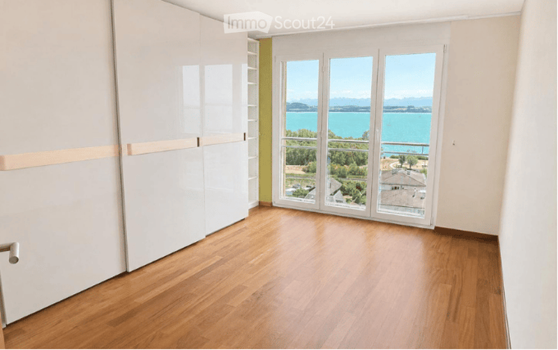 Empty room with wooden flooring, large windows offering a scenic view of a lake and surrounding landscape.