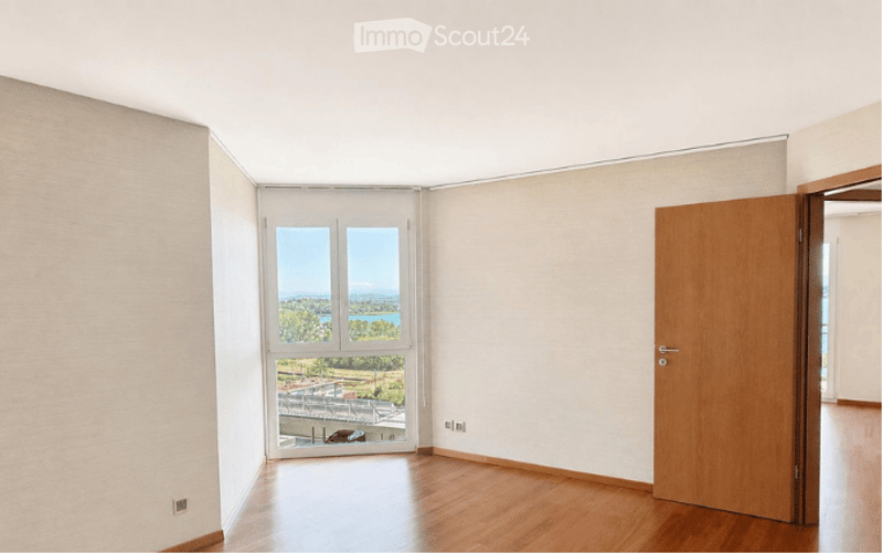Completely empty room with wooden flooring, white walls, and large windows offering a scenic view of the landscape.