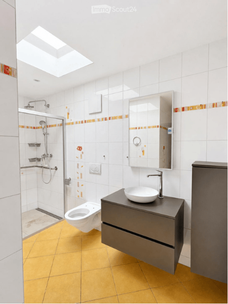 Modern bathroom with white tiles, a glass-enclosed shower, a white sink with a chrome faucet, a mirror, and a toilet. The floor is yellow tiled, and the walls feature a decorative orange stripe.