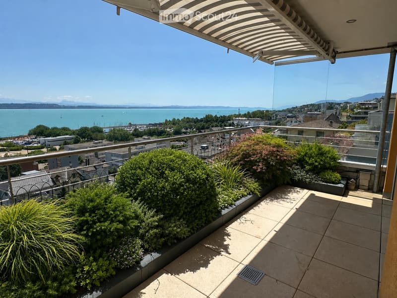 Balcony with modern glass railing, tiled floor, and lush green plants, overlooking a scenic waterfront with boats and distant mountains.