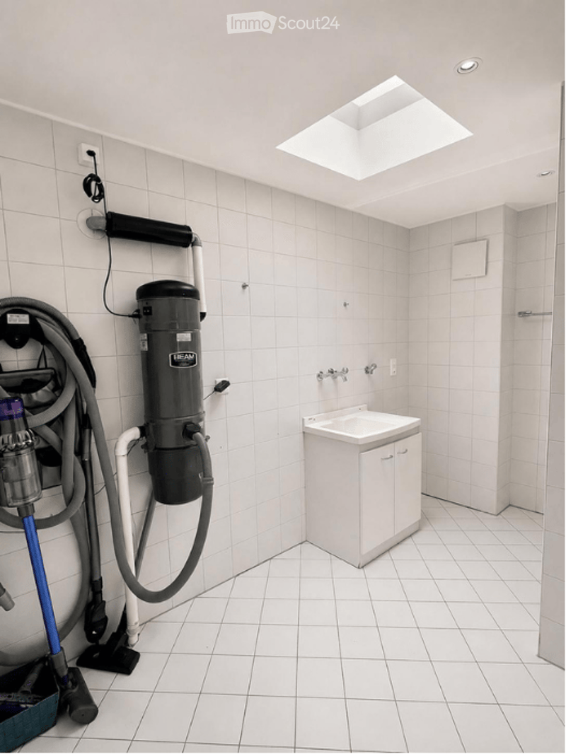 Clean, white-tiled bathroom with a sink, cabinet, and vacuum cleaner stored against the wall.