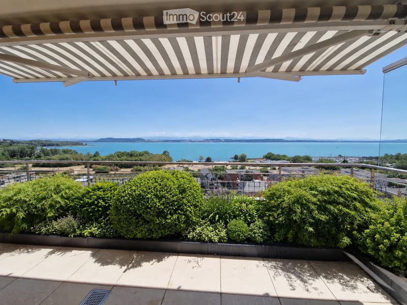 Balcony with striped awning, metal railing, and lush green shrubs, offering a scenic view of the ocean and distant hills.