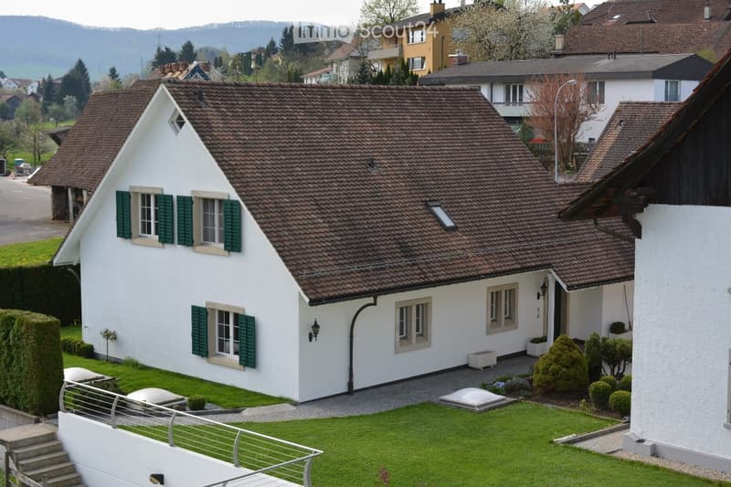 A two-story white house with a brown tiled roof, green shutters, and a well-maintained lawn in a suburban setting.