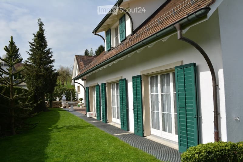 A well-maintained garden area with a neatly trimmed lawn, a large evergreen tree, and a white house with green shutters and a brown tiled roof in the background.