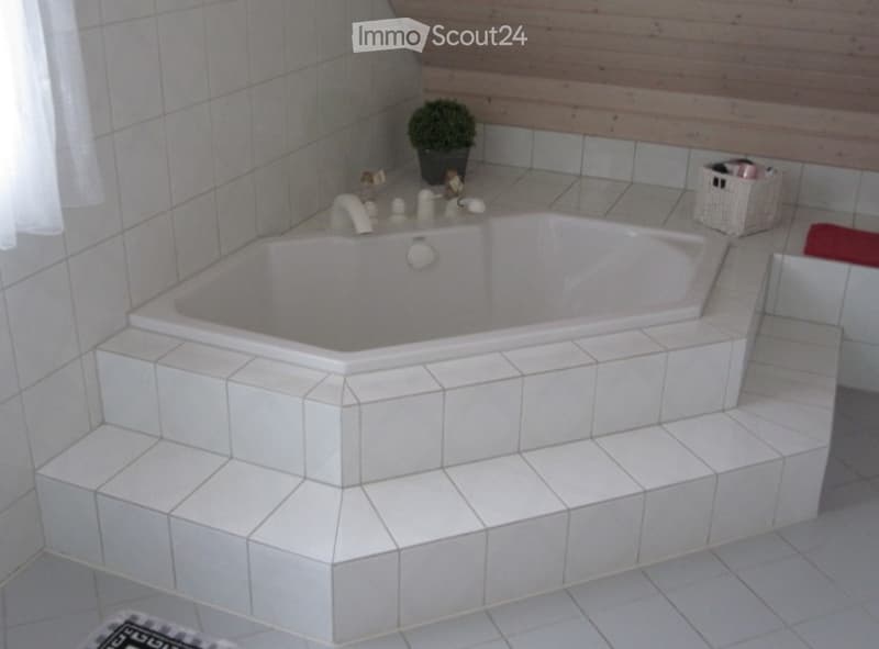 Modern bathroom with a large, corner bathtub featuring tiled steps leading into it. The walls are tiled, and there is a small potted plant and a wicker basket on the counter.
