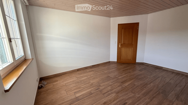 Empty room with wooden flooring, white walls, a wooden door, and a window with a wooden sill. No furniture or equipment is present.