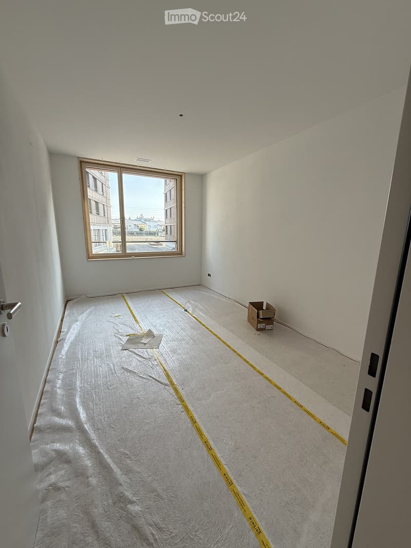 Empty room with white walls, a large window, and a covered floor with protective plastic and yellow measuring tape. A small cardboard box is visible near the window.