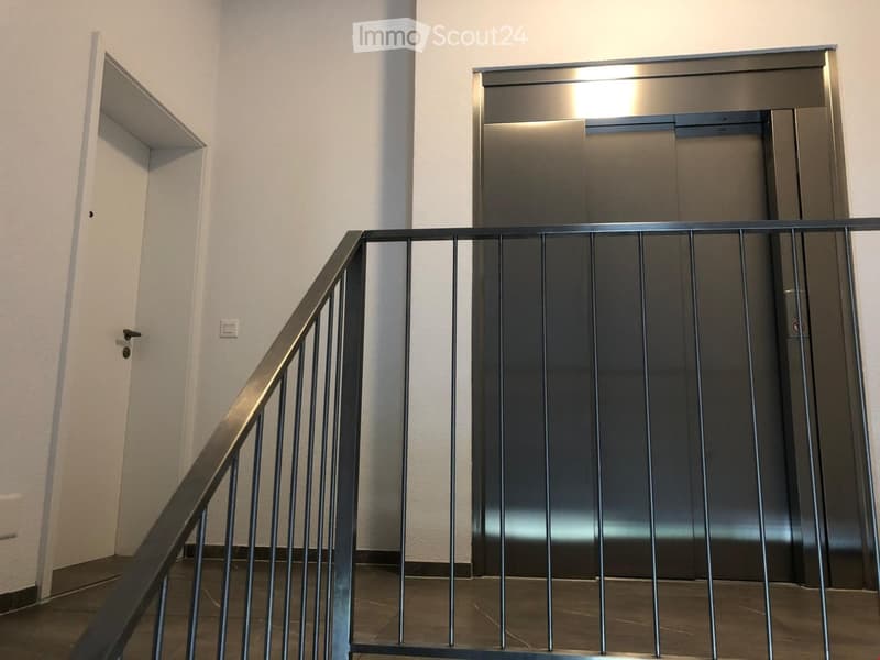 Modern hallway with a metal staircase railing, elevator doors, and a light switch on the wall.