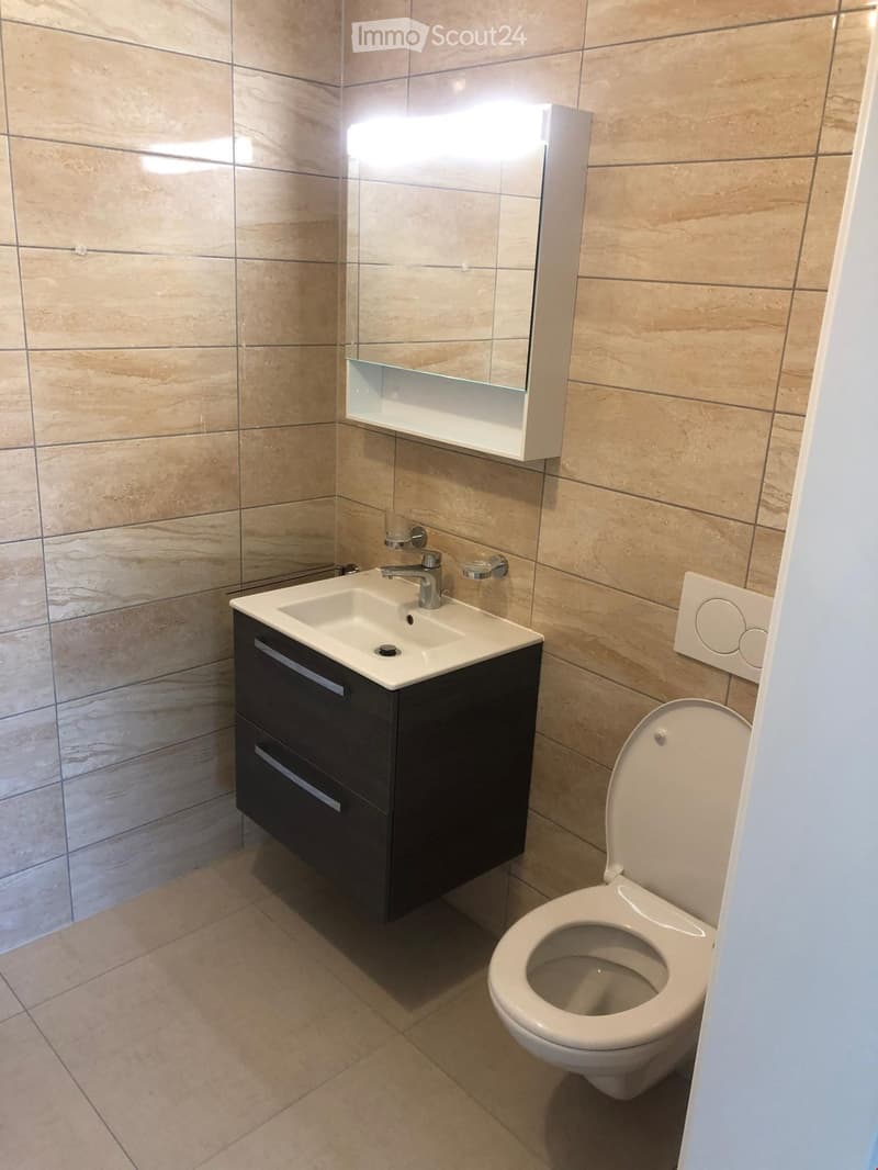 Modern bathroom with beige tiled walls and floor, a white sink with a dark wooden cabinet, a chrome faucet, a large mirror with built-in lighting, and a toilet with the lid up.