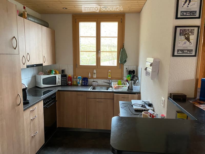 Modern kitchen with wooden cabinets, stainless steel appliances, a microwave, oven, sink, and a laptop on the counter. There are framed pictures on the wall and a window letting in natural light.