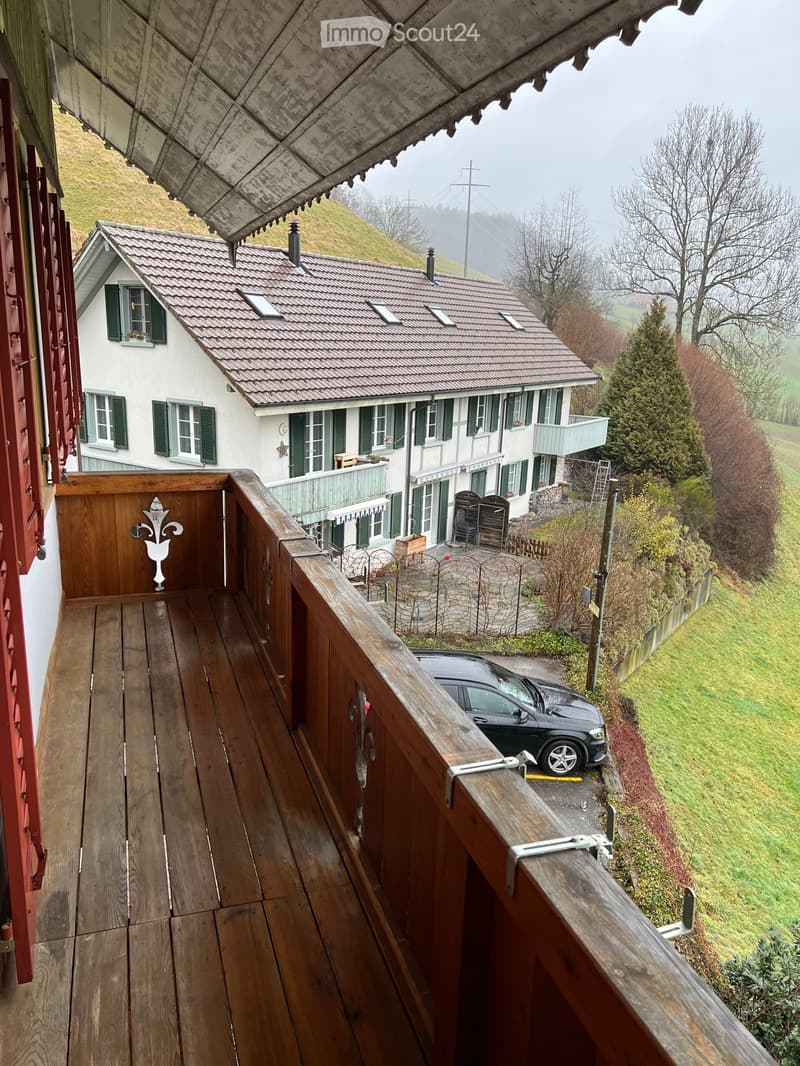 Wooden balcony overlooking a two-story house with green shutters, surrounded by a grassy hillside and trees.