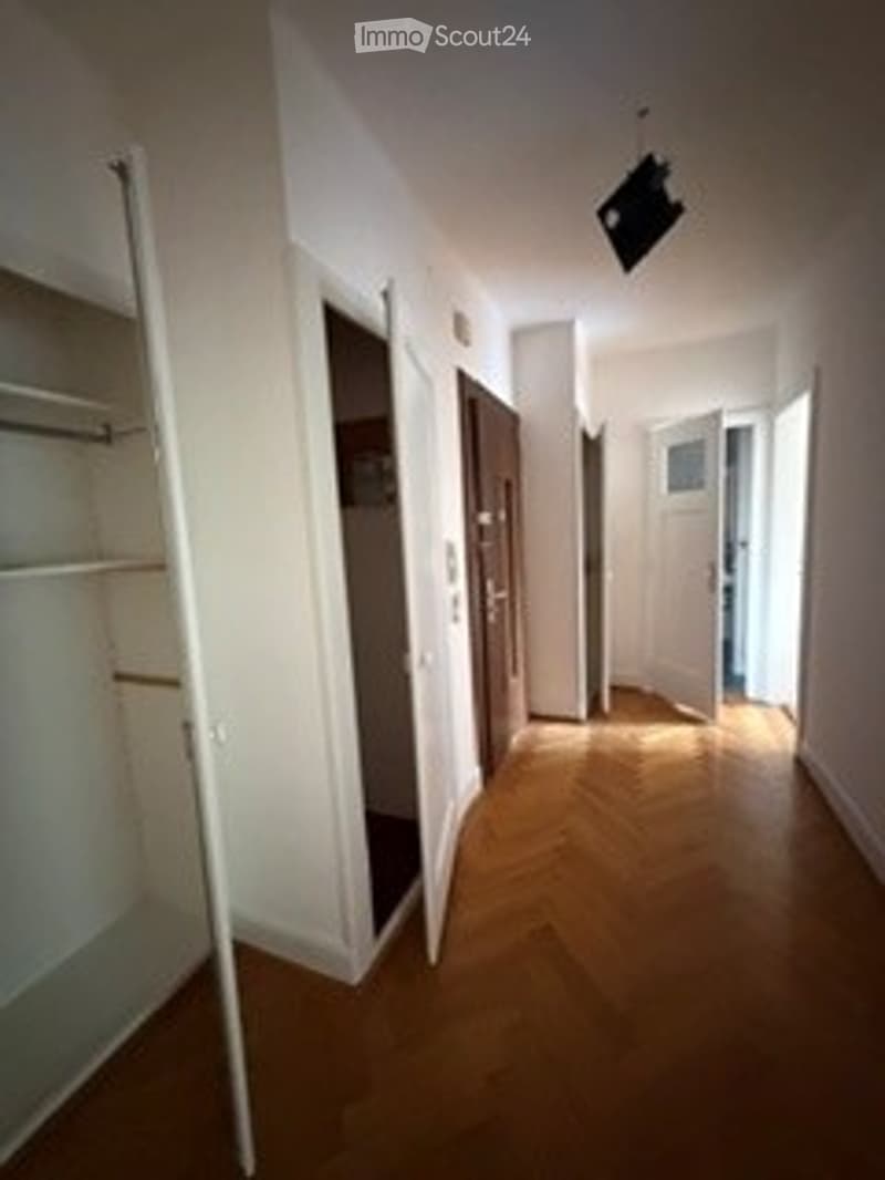 A narrow hallway with wooden flooring, white walls, and several doors leading to different rooms. The space is well-lit with natural light coming from an open doorway at the end.