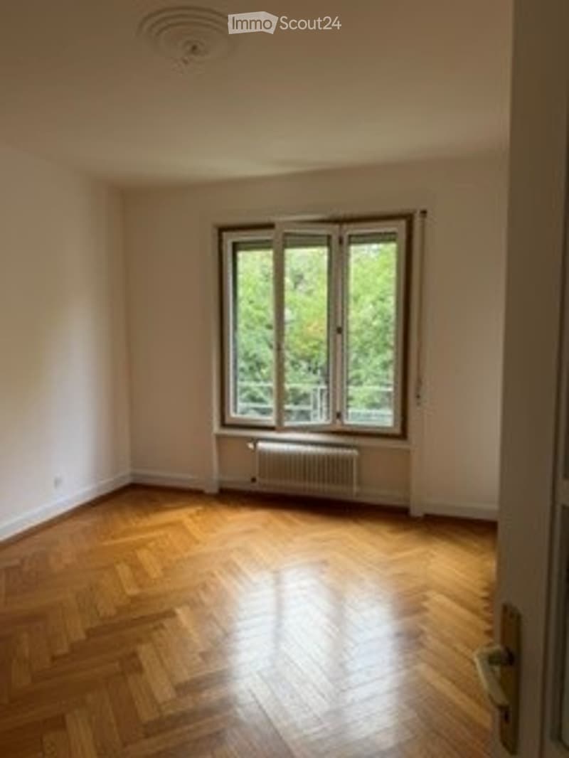 Completely empty room with wooden floor, large window, radiator, and white walls.