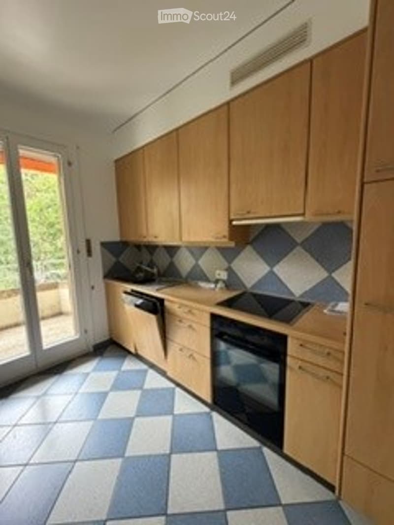 Modern kitchen with light wooden cabinets, black appliances, and a checkered tile floor. Features a dishwasher, oven, and a sink, with a sliding glass door leading to a balcony.