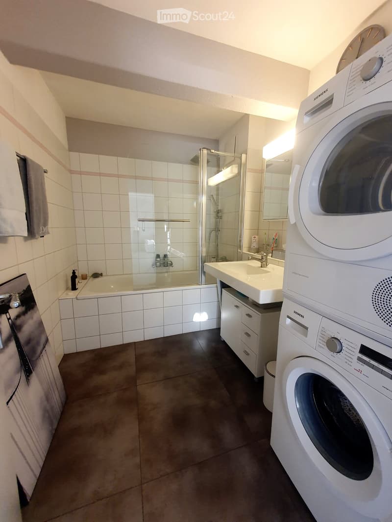 Modern bathroom with a bathtub, shower stall, sink, mirror, and a washing machine. The space features white tiled walls and a dark tiled floor.