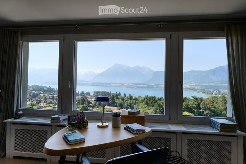 Bright room with large windows offering a scenic mountain and lake view. Features a round wooden table with books, a lamp, and a flower arrangement on the windowsill.