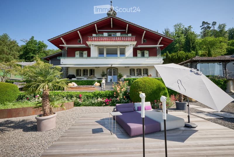 A large, elegant garden area with a wooden deck, a purple outdoor sofa, a small table, and a large umbrella. The background features a red and white house with multiple balconies and lush greenery.