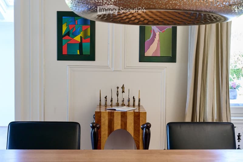 Elegant room with a wooden table, black leather chairs, a decorative side table with small sculptures, framed artwork, and a large window with curtains.