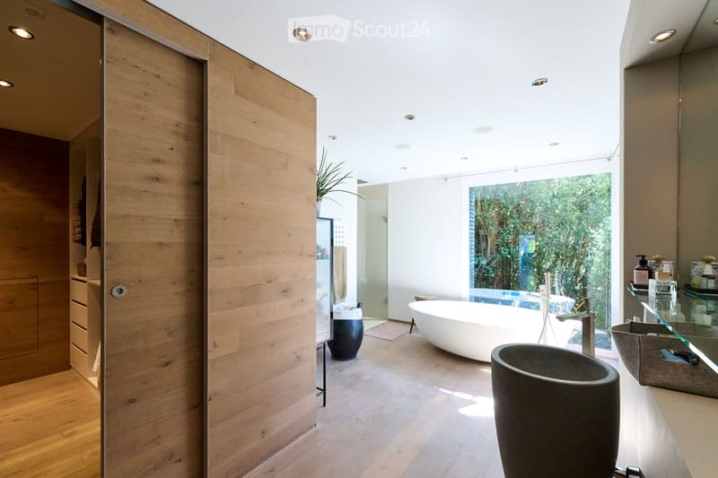 Modern bathroom with a freestanding bathtub, sink, and mirror. Features wooden flooring, large windows with a view of greenery, and built-in storage cabinets.