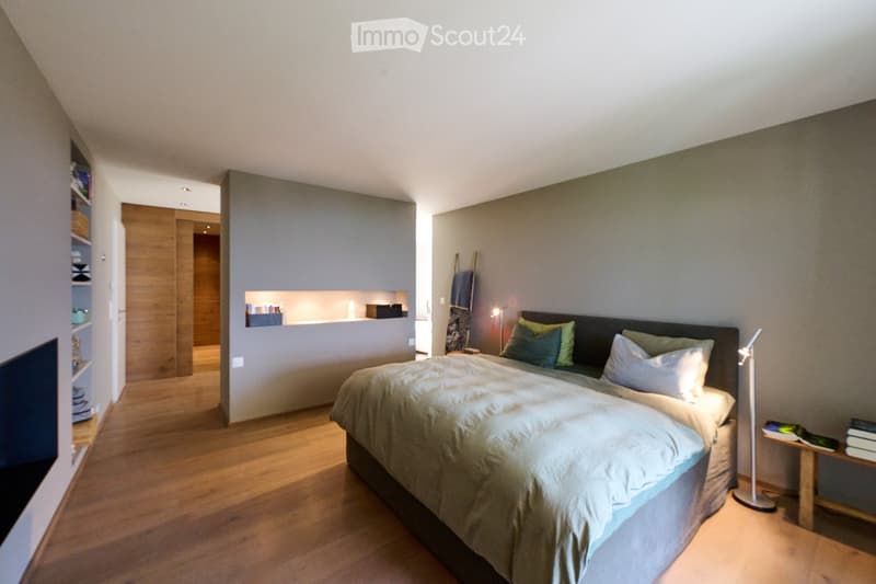Modern bedroom with a large bed, nightstand, lamp, and built-in shelving unit. The room features wooden flooring and neutral-colored walls.