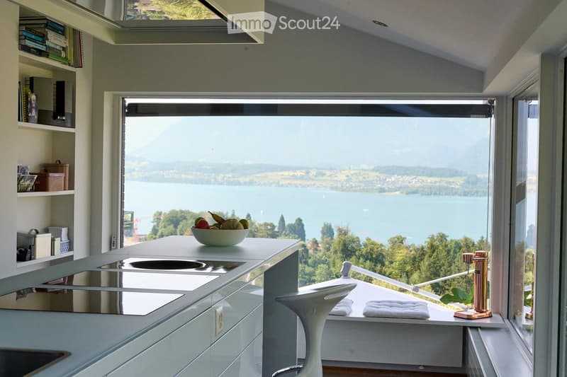 Modern kitchen with sleek white countertops, built-in sink, and a large window offering a scenic lake view. Includes a white stool, a bowl of fruit, and shelves with books and decorative items.