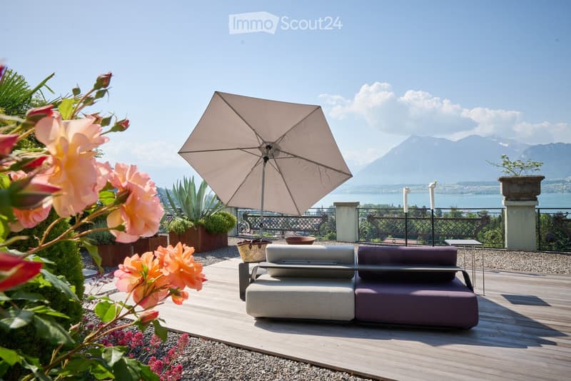 A sunny outdoor patio with a wooden deck, a grey sofa, a large umbrella, a small table, and various potted plants, overlooking a scenic lake and mountains.