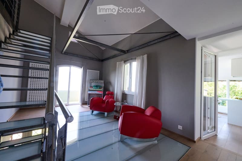 Modern living room with a glass floor, red chairs, a glass coffee table, and large windows with white curtains. A staircase leads to an upper level.