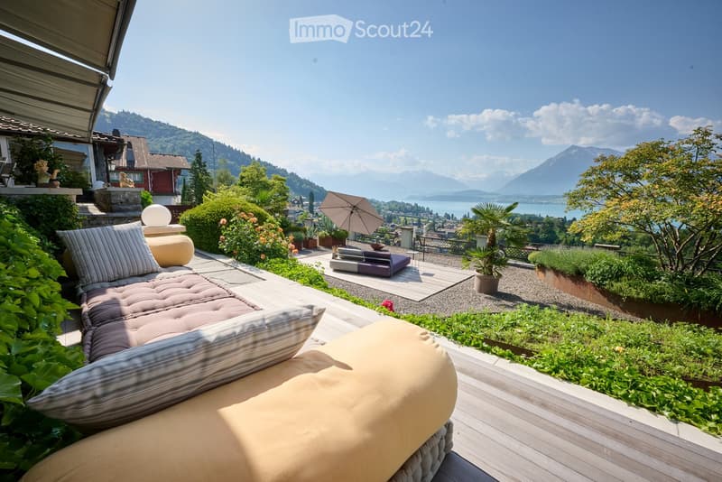 A well-maintained garden with a comfortable sofa, striped cushions, and a small umbrella, overlooking a scenic lake and mountains.