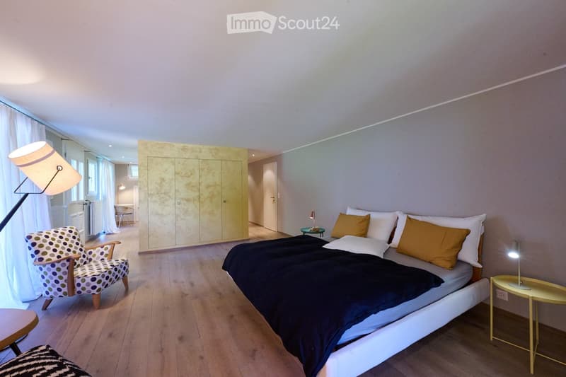Modern bedroom with a large bed, patterned chair, wardrobe, side table, and floor lamp. The room features wooden flooring and large windows with white curtains.