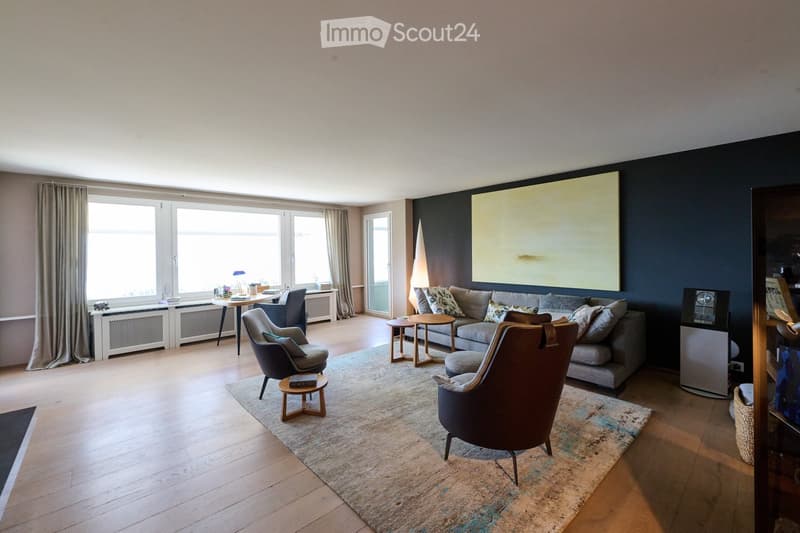 Modern living room with light wooden flooring, large windows with curtains, a grey sofa with patterned cushions, two armchairs, a coffee table, a side table, and a large abstract painting on the wall.