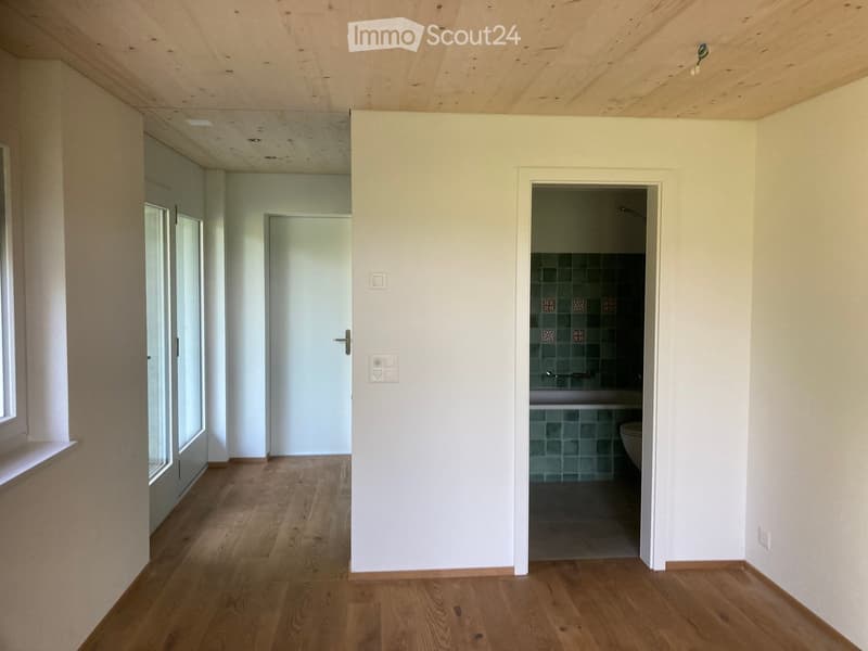 Empty hallway with wooden flooring, white walls, and a partially visible bathroom with green tiles and a toilet.