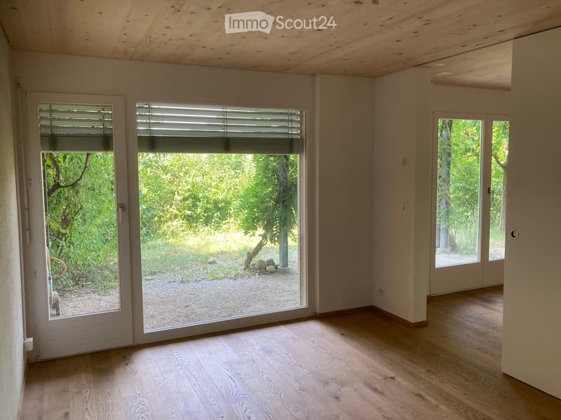 Empty room with wooden flooring, large windows, and sliding doors leading to a garden area.