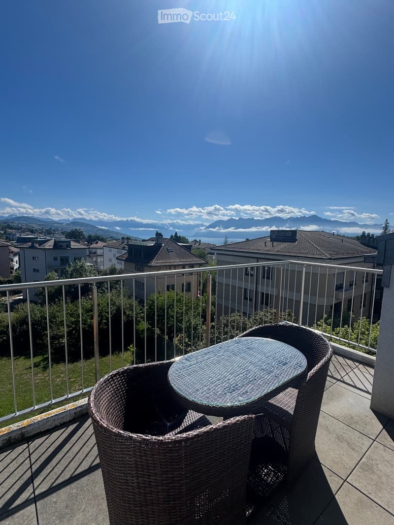Balcony with a glass table and two wicker chairs, overlooking a scenic view of mountains and a lake.