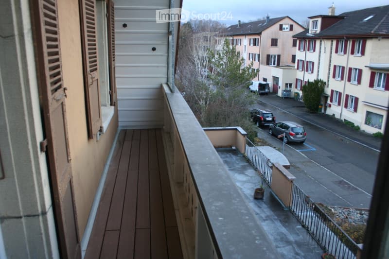 An exterior view of a balcony with wooden floor in front of a building with multiple windows and some trees