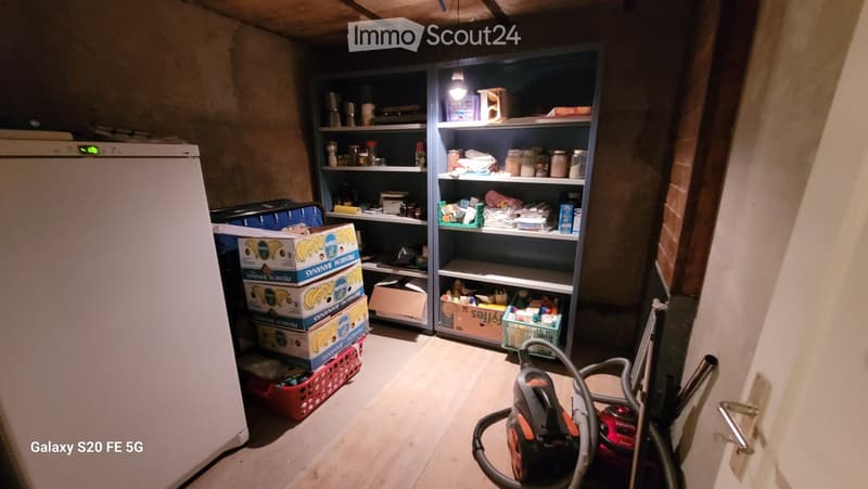 A storage room with a refrigerator, shelves filled with various items, boxes stacked on the floor, and a vacuum cleaner near the door.