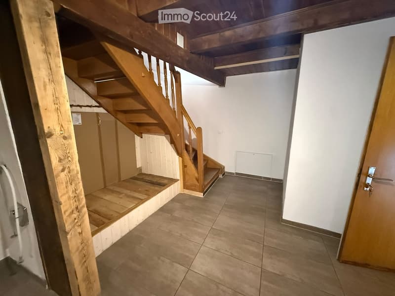 Wooden staircase with a handrail leads to an upper level, flanked by white walls and tiled flooring.