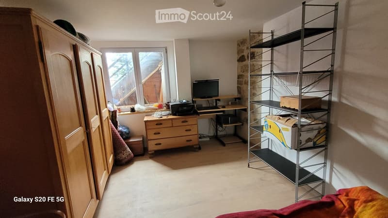 A furnished room with a desk, chair, computer, printer, wardrobe, shelf, and mirror. The room has a window with a view of a wooden structure outside.