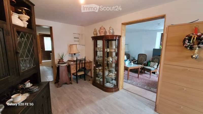 A well-furnished room with a wooden floor, a large cabinet with glass doors, a display cabinet, a small table with a lamp, and a doorway leading to another room with a sofa and chairs.