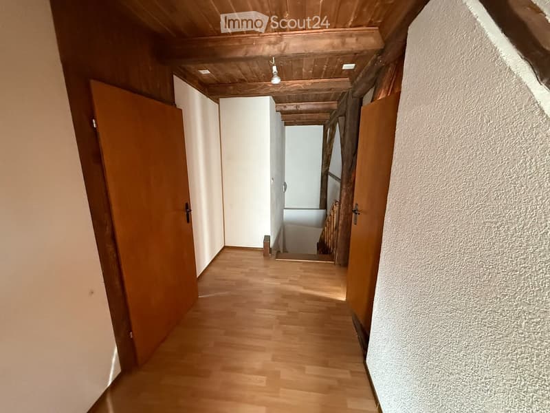 A narrow hallway with wooden flooring, wooden doors on both sides, and a visible staircase leading upwards.