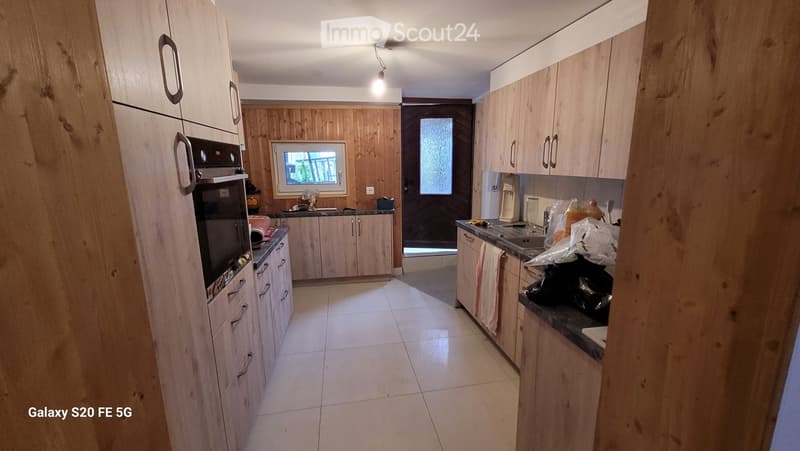 A modern kitchen with light wooden cabinets, a tiled floor, and a central island. The kitchen features an oven, a sink, and a refrigerator, with a window and a door leading outside.