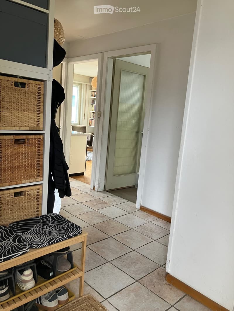 Hallway with tiled floor, wooden bench with shoes, coat rack with coats and baskets, and a door leading to another room.