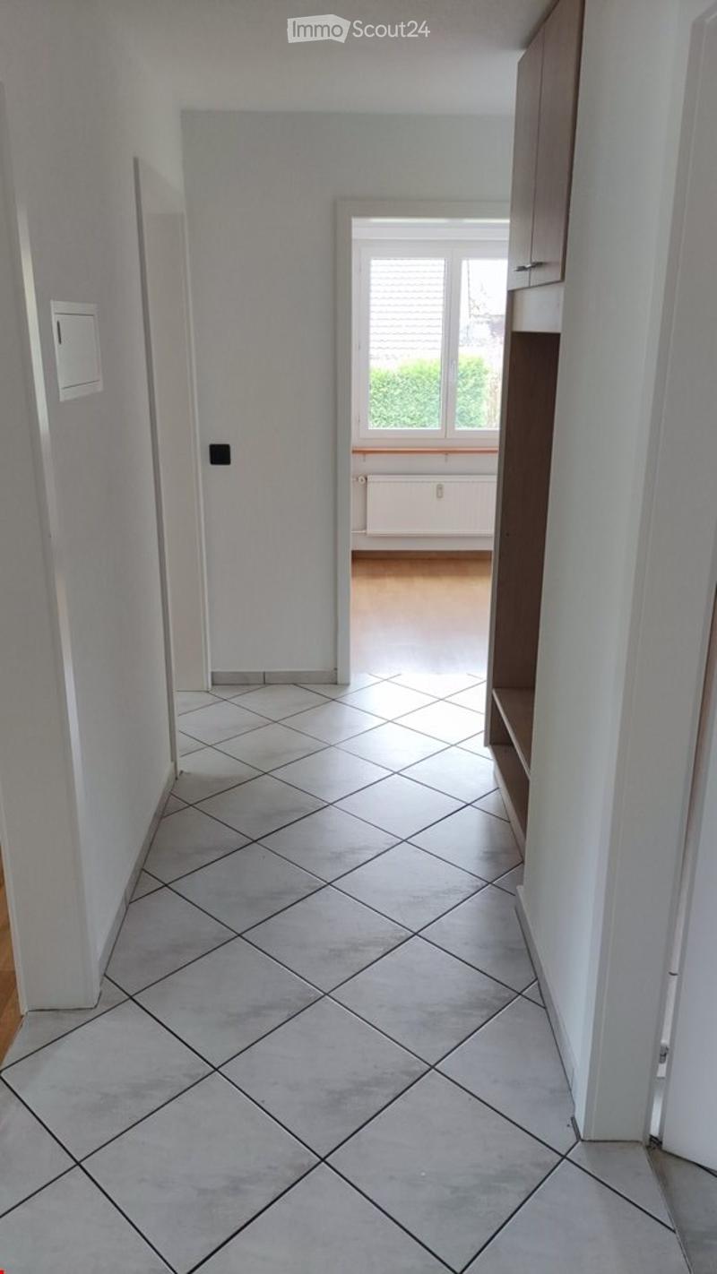 Empty hallway with tiled floor and white walls, leading to a room with wooden flooring and a window.