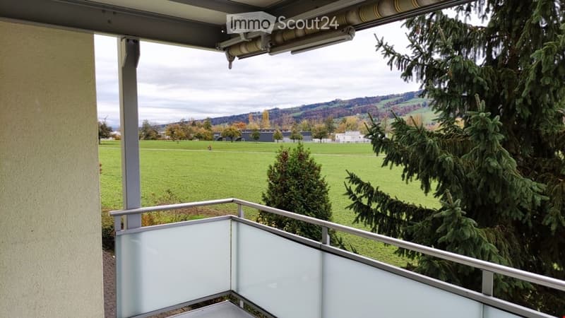 Balcony with a view of a green field, trees, and distant hills under a cloudy sky.